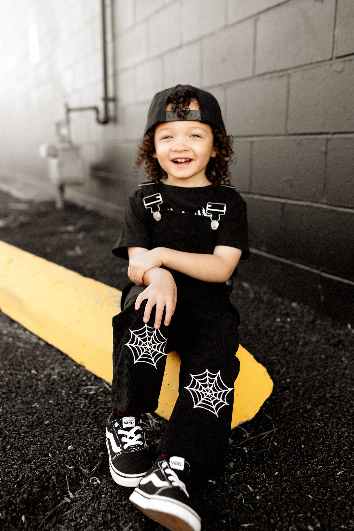 Black kids’ overalls with front straps and white spiderweb patches on legs.