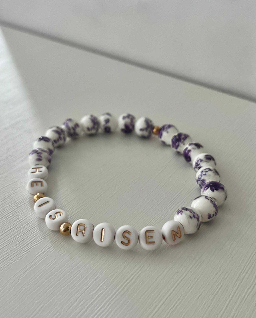 Purple-speckled bead bracelet with white letter beads spelling “HE IS RISEN”