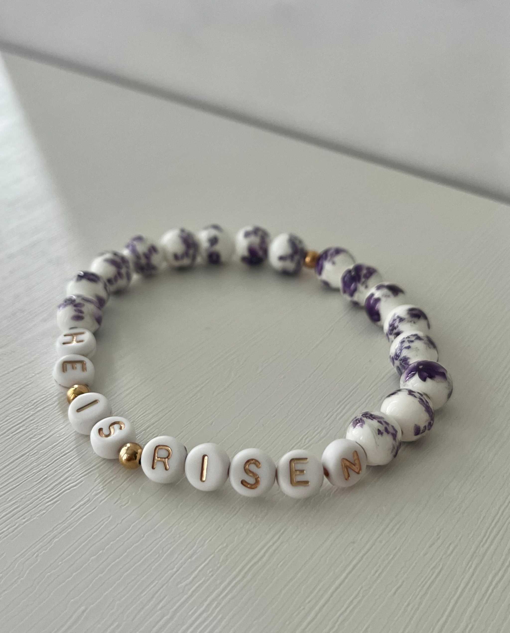 Purple-speckled bead bracelet with white letter beads spelling “HE IS RISEN”