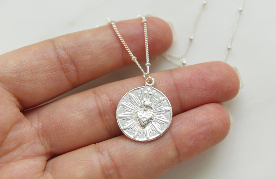 Silver round sacred heart coin pendant necklace held in hand on delicate chain.