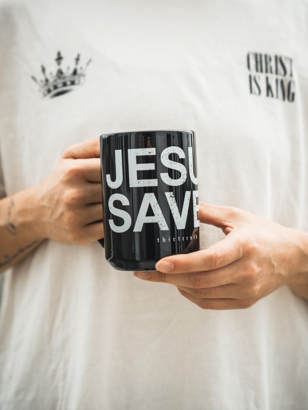 Black mug held in hands, 'JESU SAV' text with crown graphic and 'thirteenth'