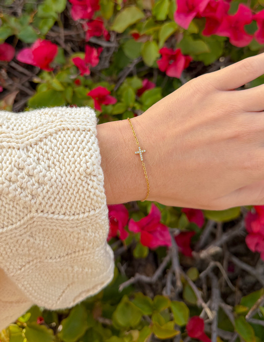 Gold cross charm bracelet worn on wrist, shown with sweater sleeve.