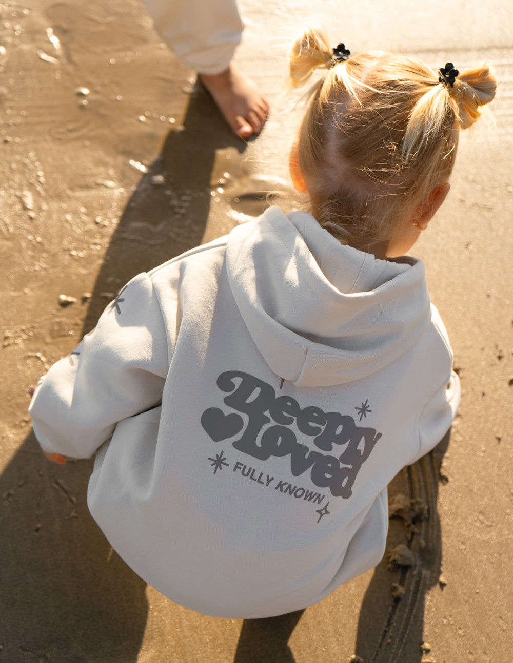 Kids hoodie off-white with “Sleepy Loved” logo on back, worn on outdoor ground