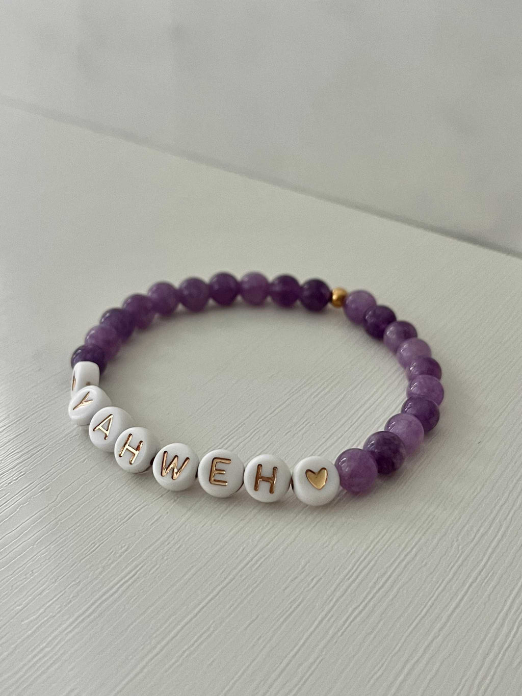 Purple beaded stretch bracelet with white letter charms spelling YAHWEH and heart details.