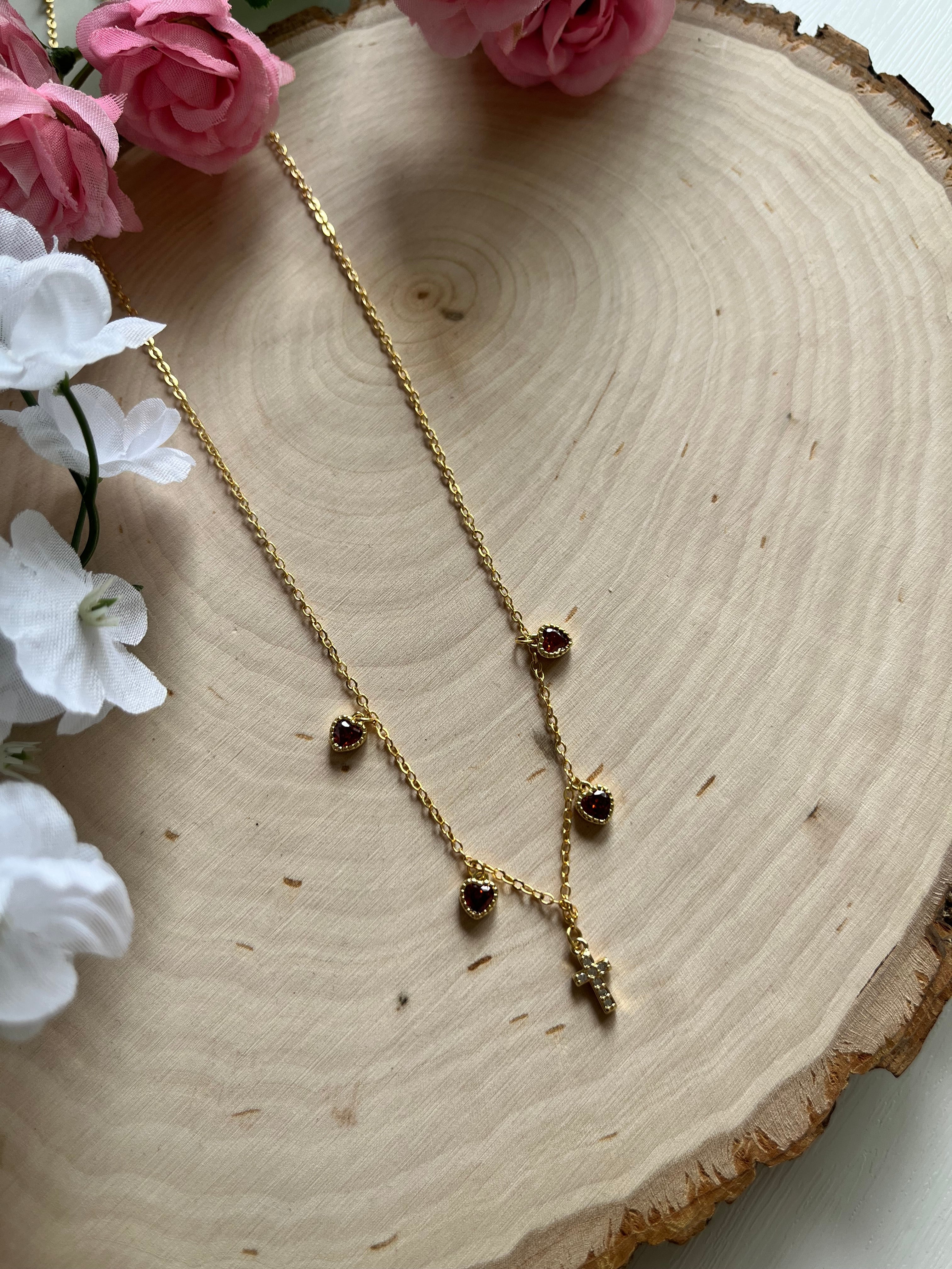 Gold charm necklace with red heart stones and gold cross charm, angled close-up