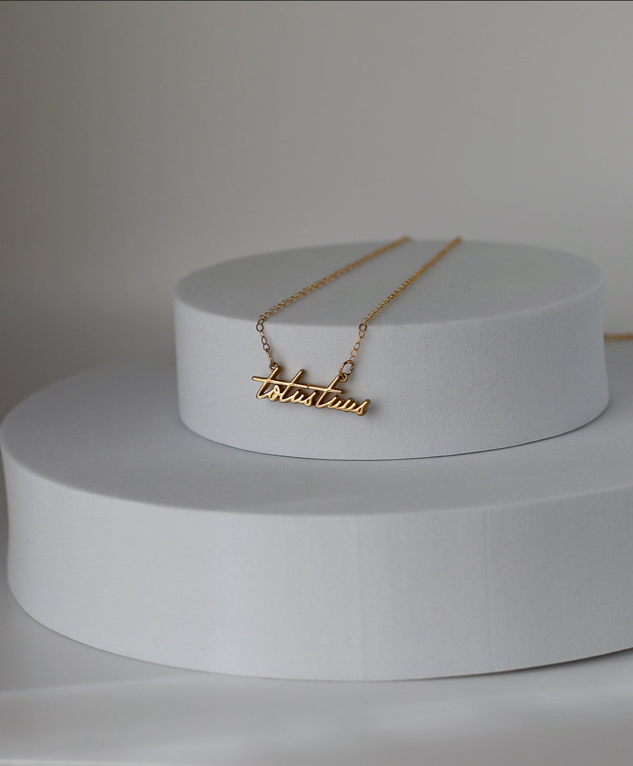 Gold-tone Totus Tuus pendant necklace on a display stand