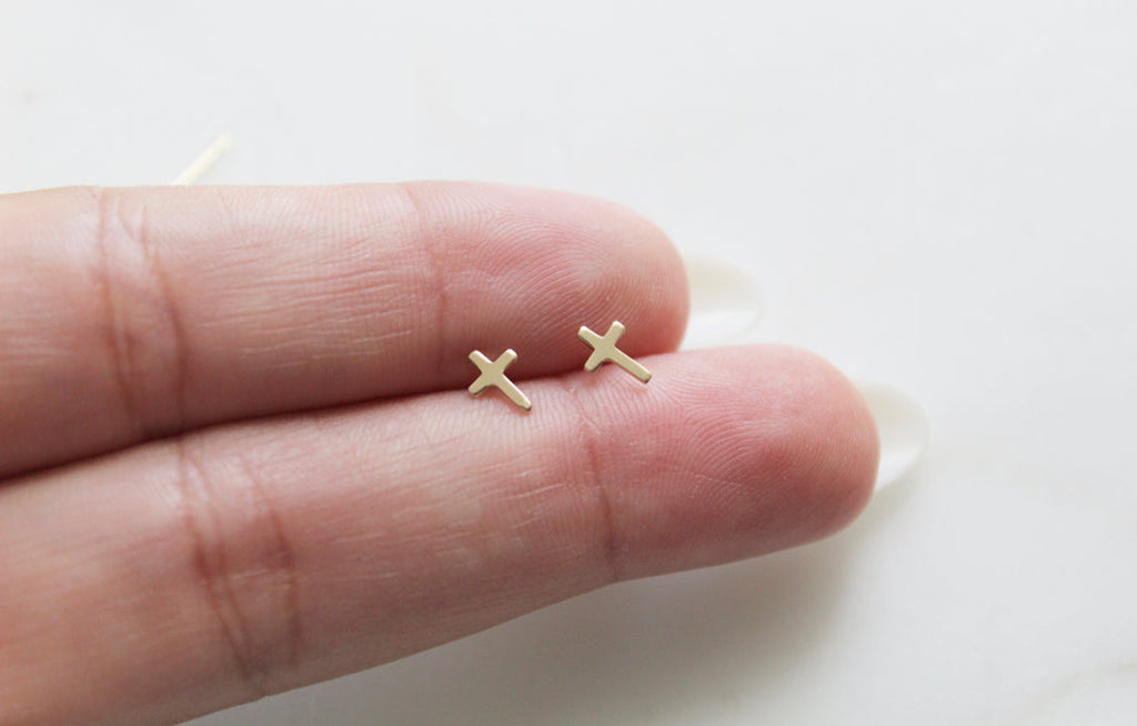 Gold-tone cross stud earrings shown on a hand, two earrings visible.