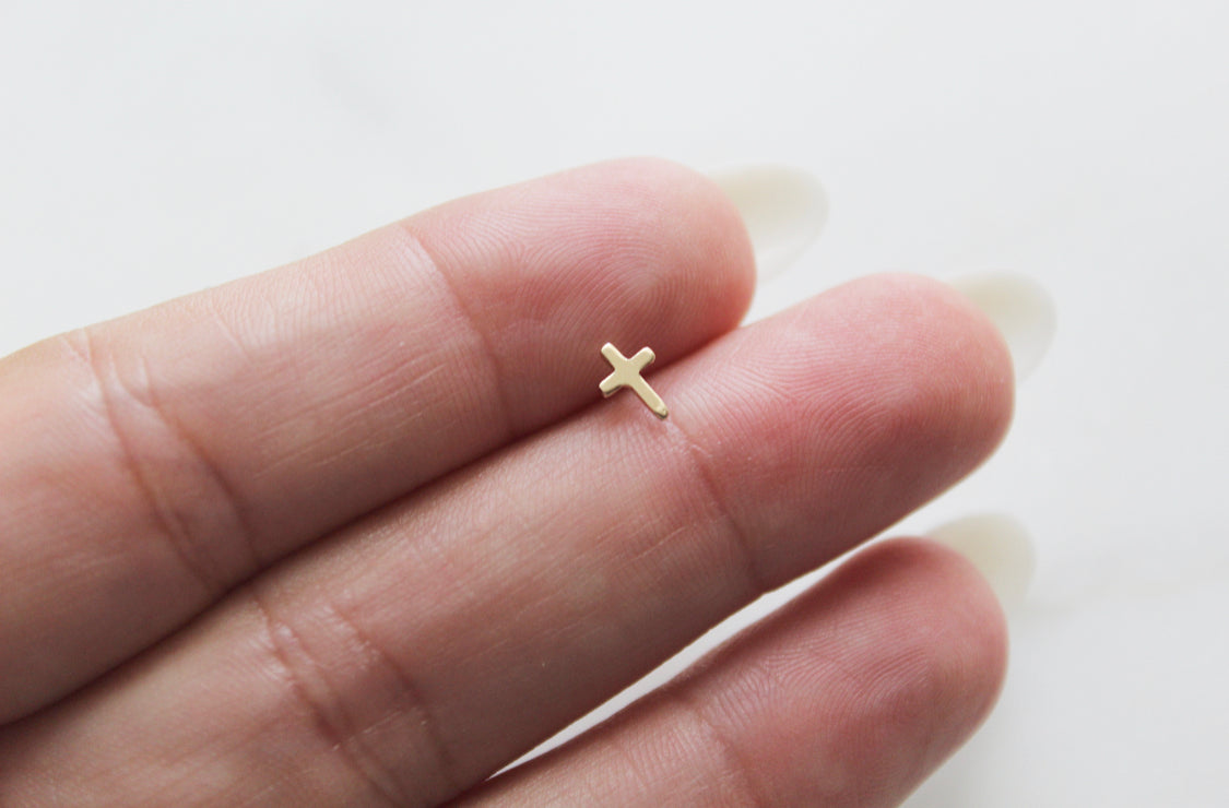 Gold-tone cross stud earring shown on a hand, front view.