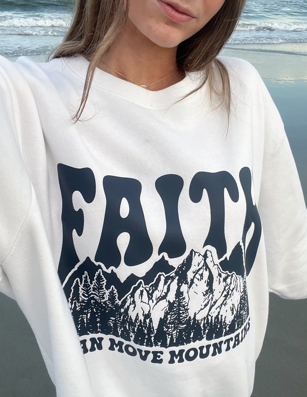 Close-up of white crewneck sweatshirt showing dark “Faith” mountain graphic across chest