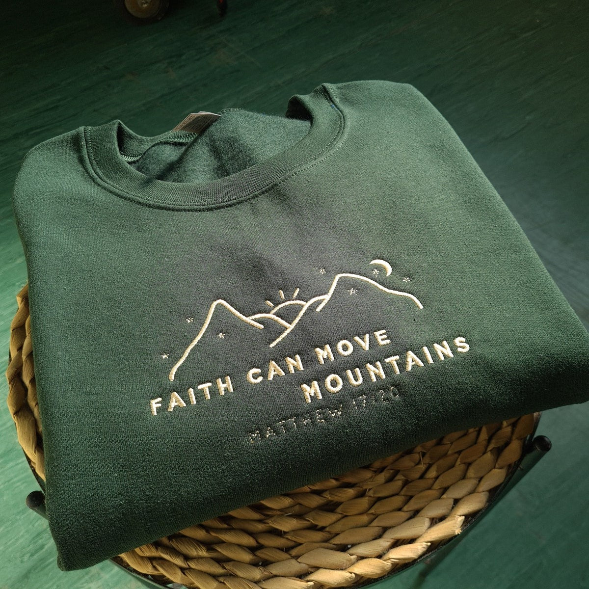 Dark green crewneck sweatshirt, folded, with embroidered “Faith Can Move Mountains” chest design