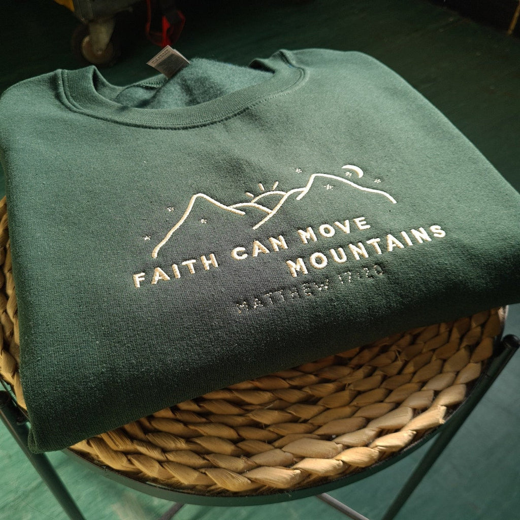 Dark green crewneck sweatshirt, folded on a basket, with embroidered faith and mountains design