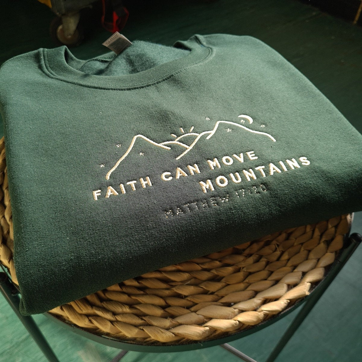Dark green crewneck sweatshirt, folded on a basket, with embroidered faith and mountains design