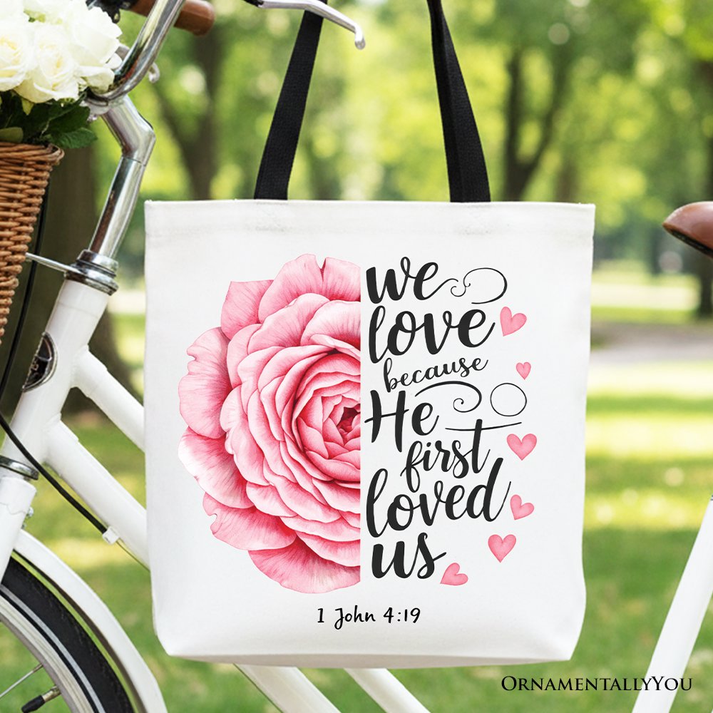 White tote bag with pink rose graphic, '1 John 4:19' text, dark handles