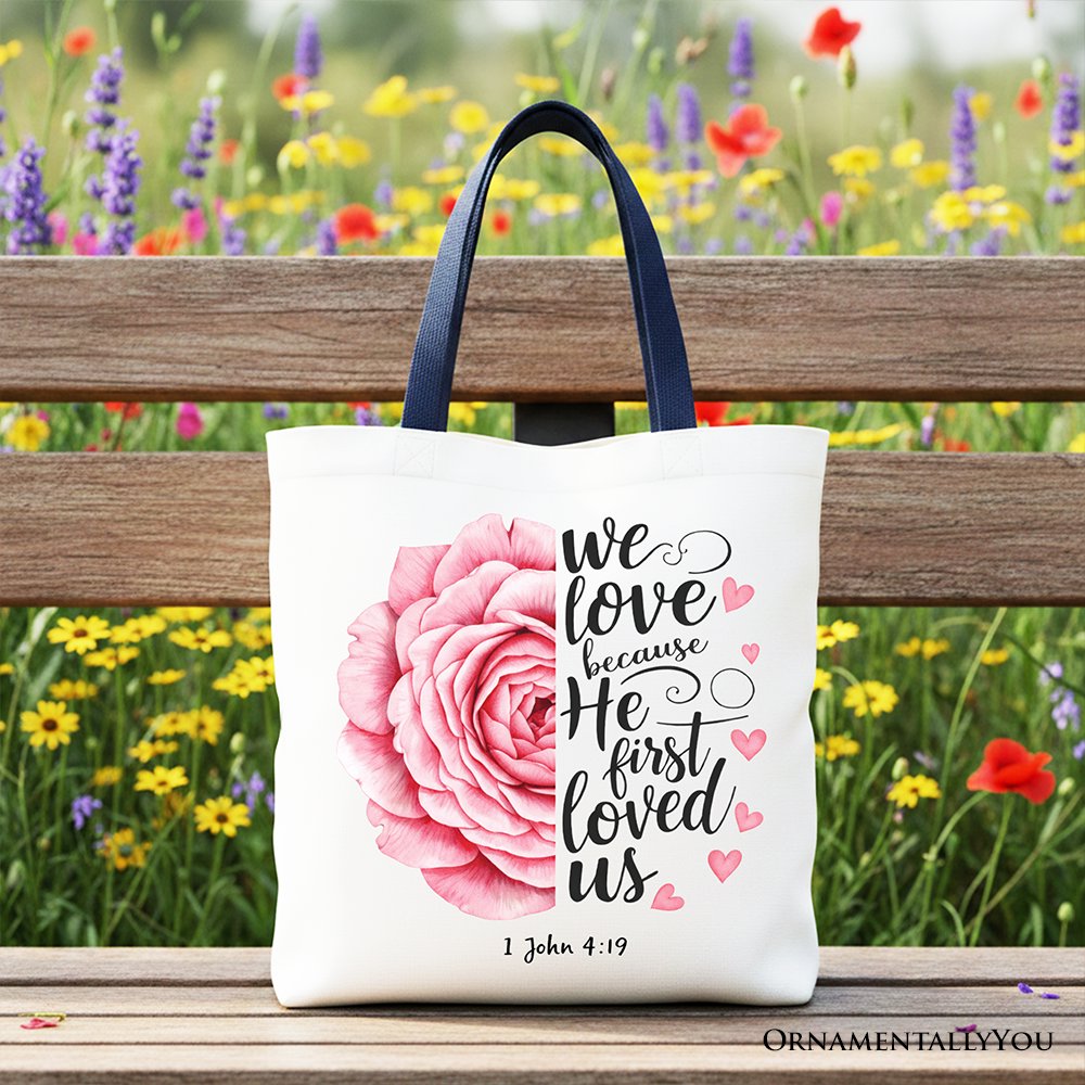White tote bag with pink rose graphic, '1 John 4:19' text, dark handles