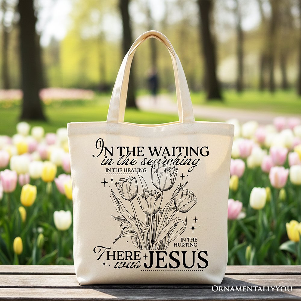 Cream tote bag with floral scripture line art and black handles, front view on bench