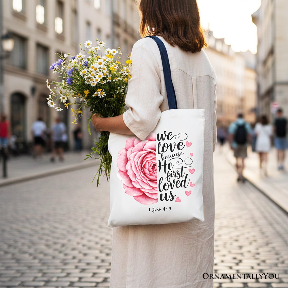 White tote bag with pink rose and '1 John 4:19' text, dark blue handles