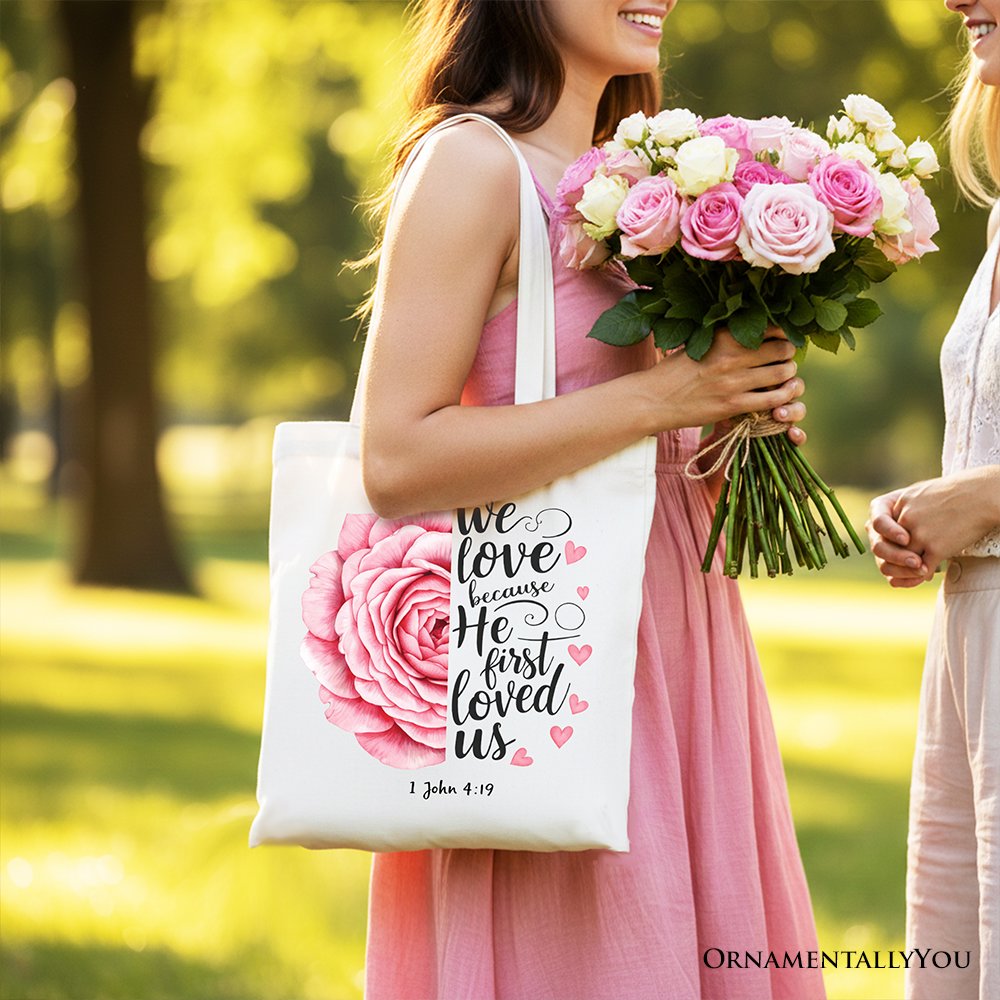 White tote bag with pink rose graphic and '1 John 4:19' text