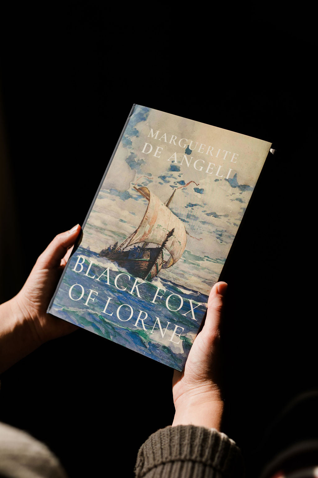 Hardcover novel held in hands, blue sky cover with sailboat illustration