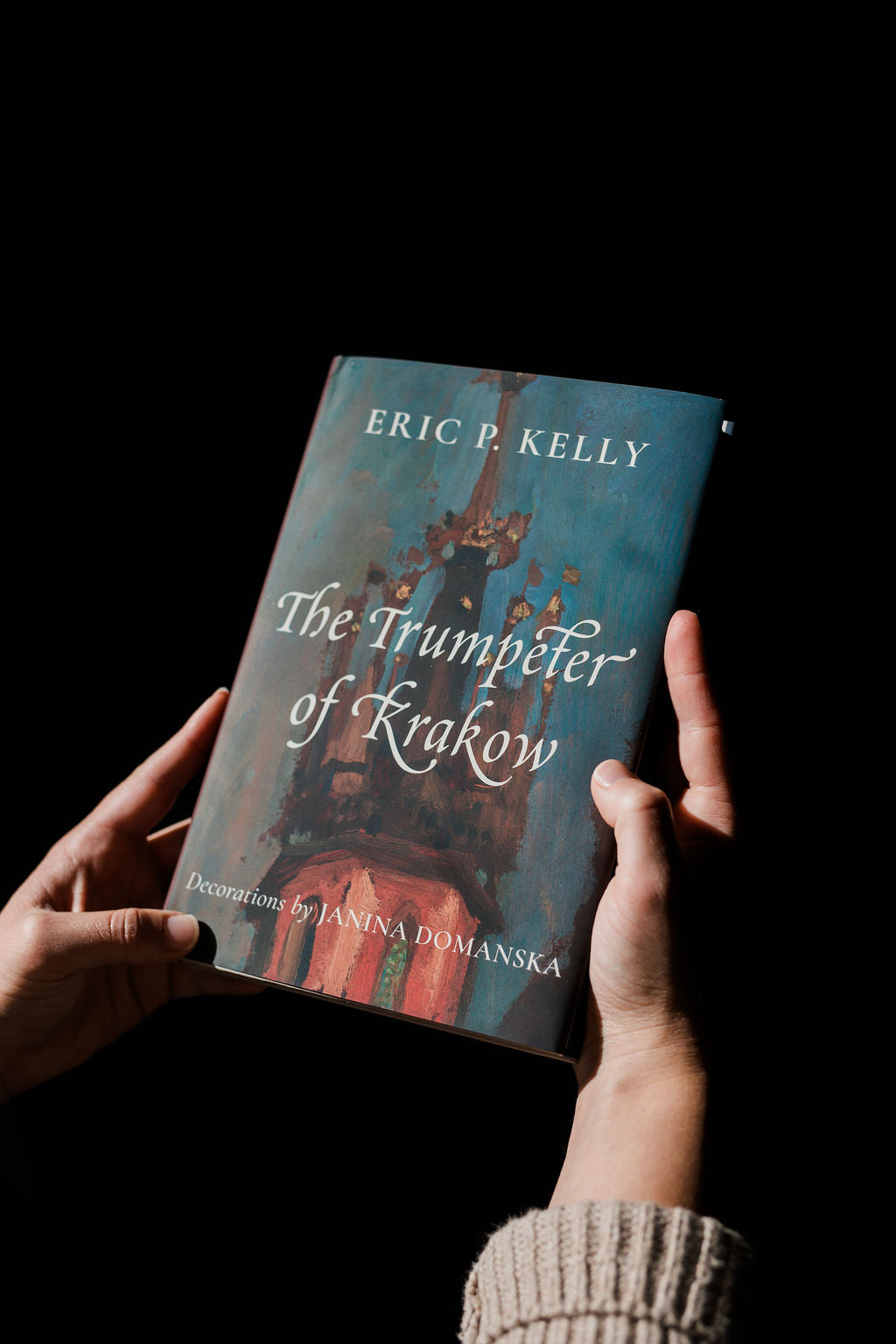 Hardcover book front cover held in hands with blue art background and ornate title type.