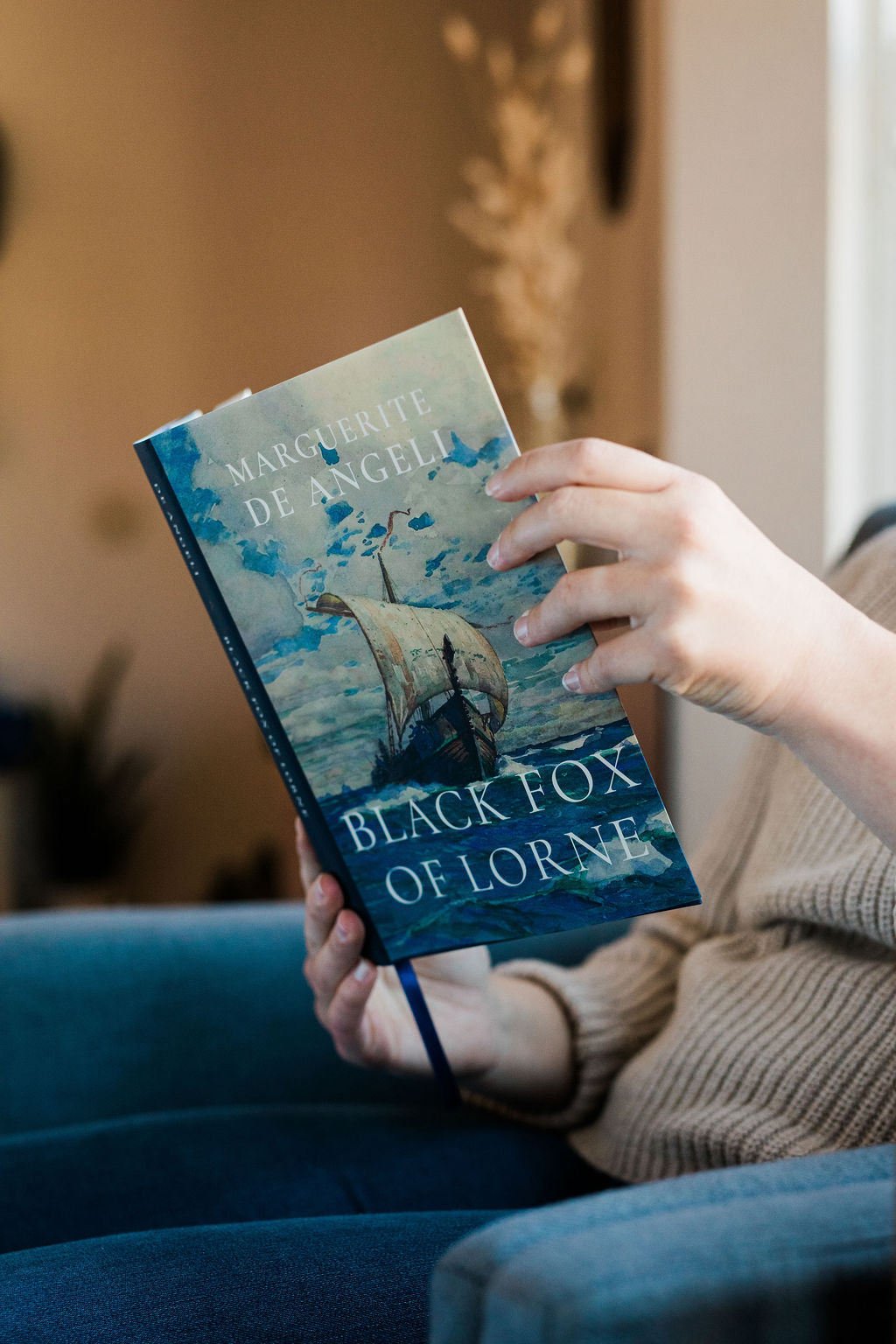 Hardcover novel held above a couch, blue sailboat cover with title lettering