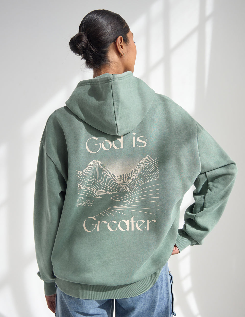 Sage green unisex hoodie from the back with “God is Greater” mountain graphic