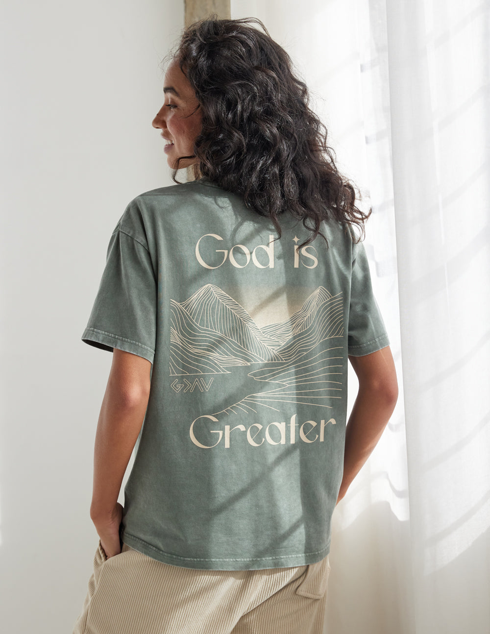Unisex graphic tee, back view in sage green with “God is Greater” mountain line art