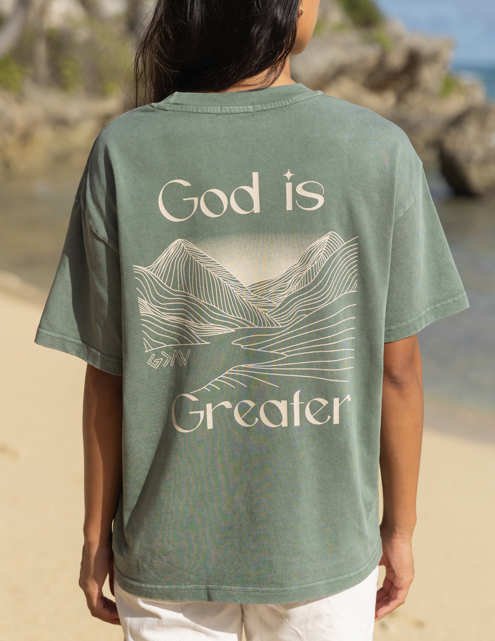 Unisex graphic tee, back view in sage green with “God is Greater” mountain line art