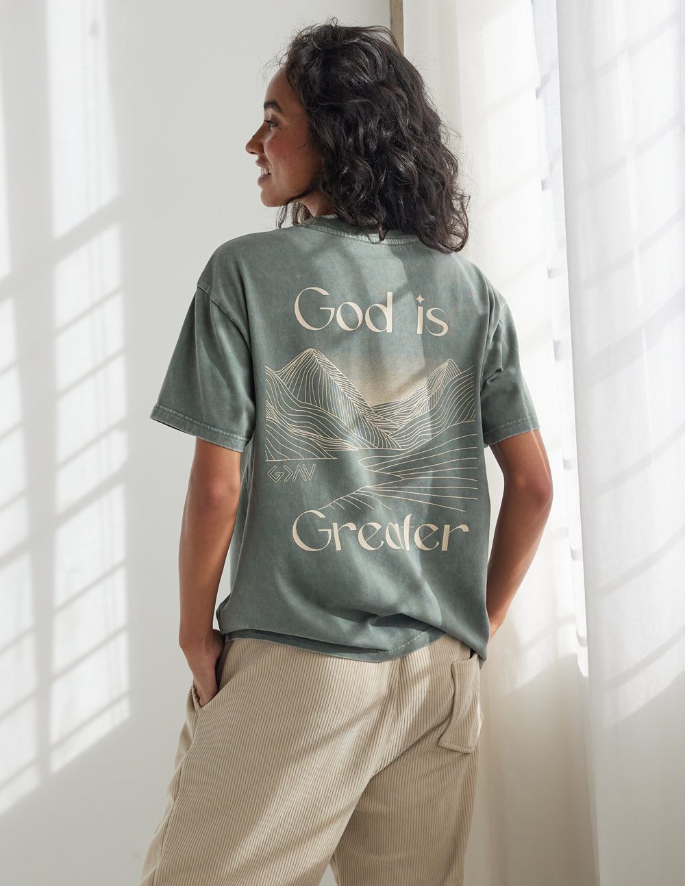 Unisex graphic tee, back view in sage green with “God is Greater” mountain line art