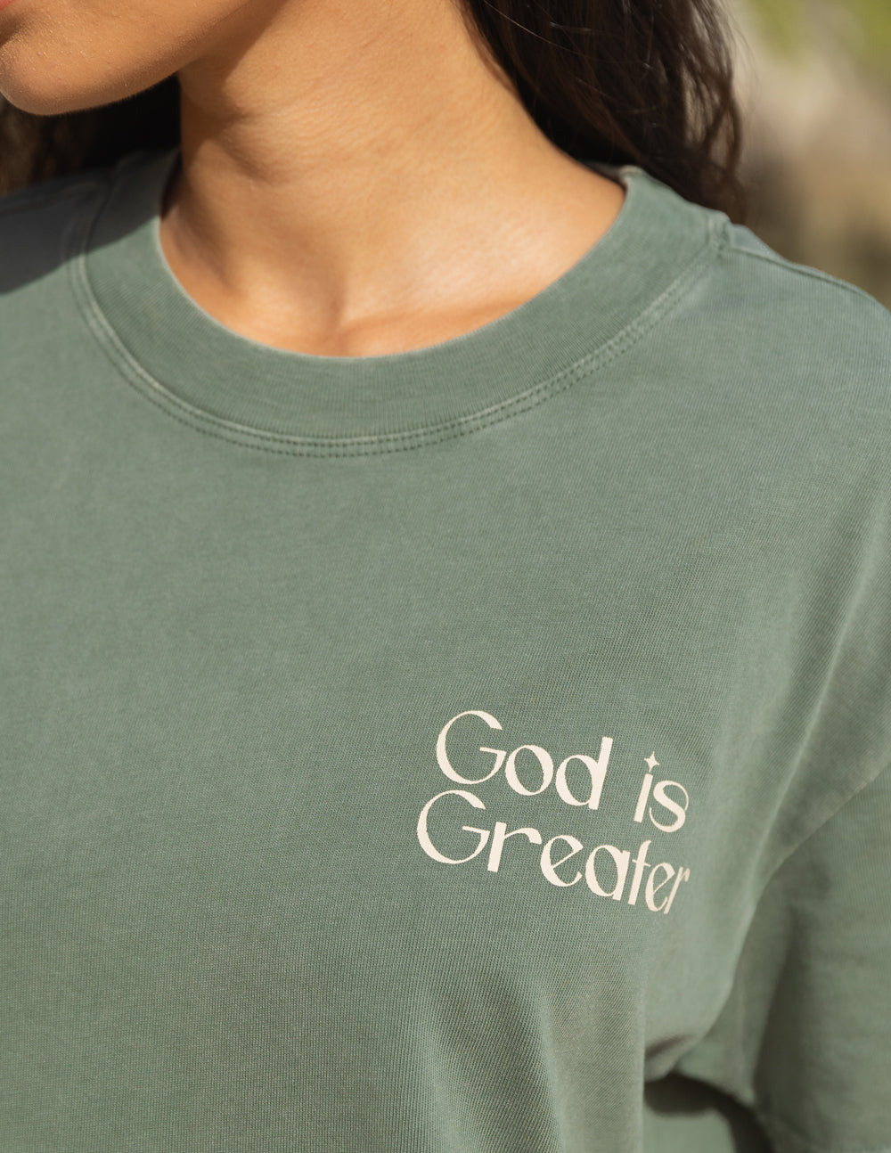 Unisex graphic tee in sage green with “God is Greater” text logo on the chest