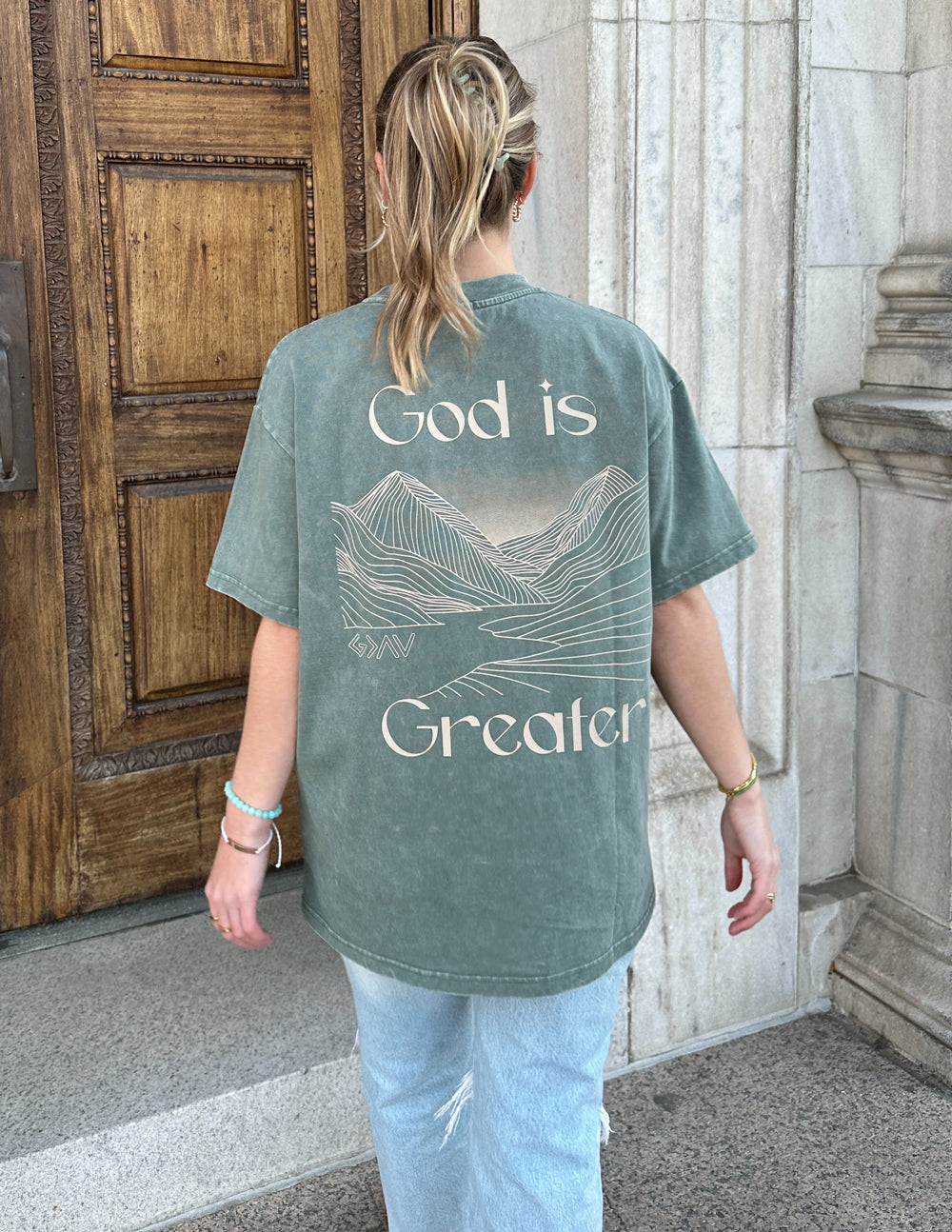 Unisex graphic tee, back view in sage green with large “God is Greater” mountain line art
