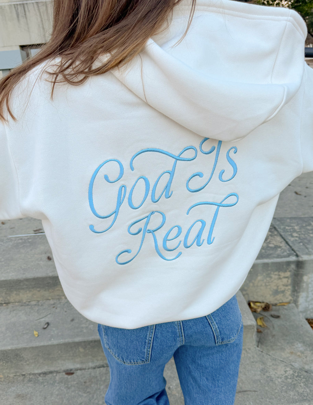 Unisex off-white hoodie, back view, large blue “God Is Real” text on hood