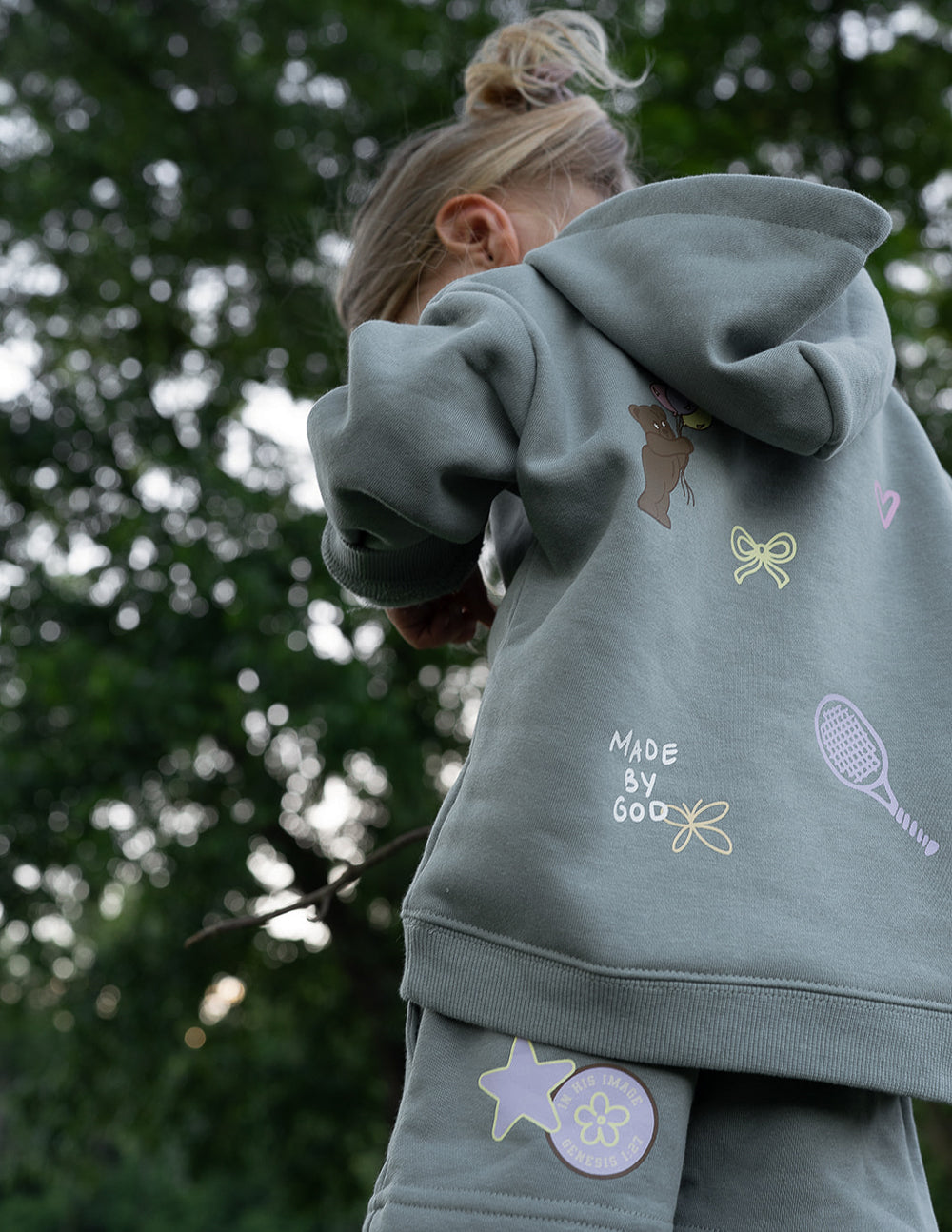 Kids sage green hoodie back print with "Made by God" and icon graphics.