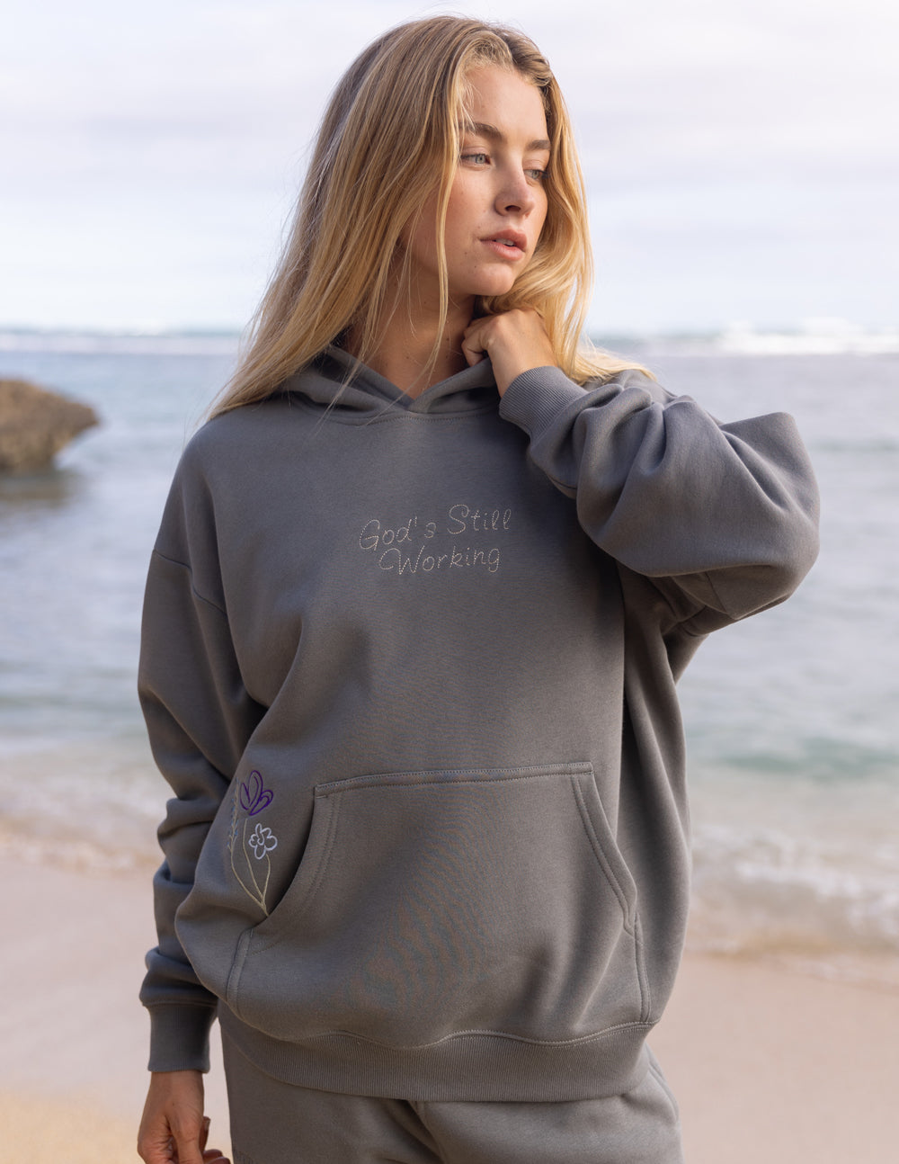 Gray unisex hoodie with front hood and small “God’s Still Working” chest text.