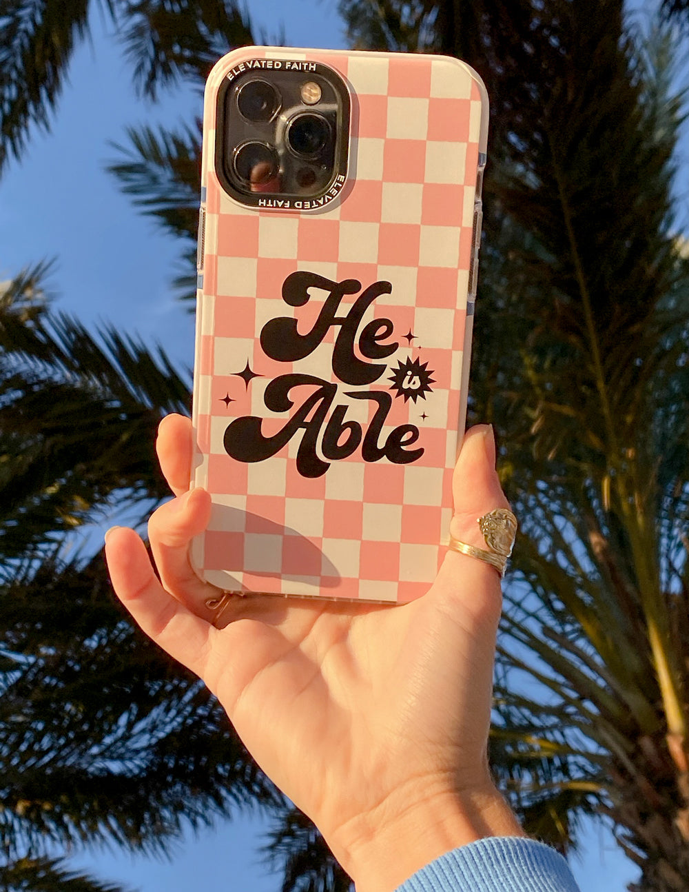 Phone case in hand, pink checkered design with 'He is Able' lettering
