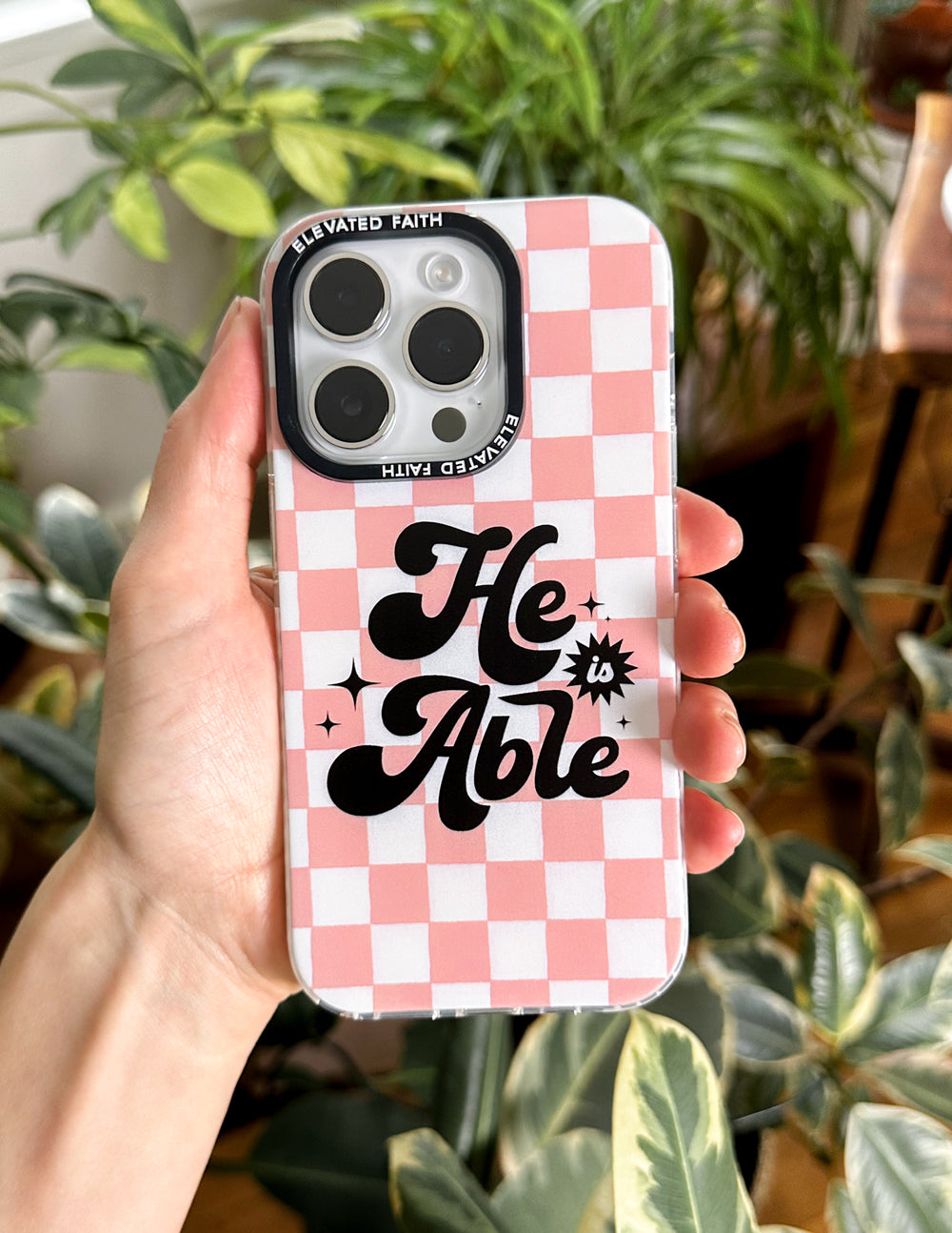 Pink checkered phone case with 'He is Able' text, close-up in hand