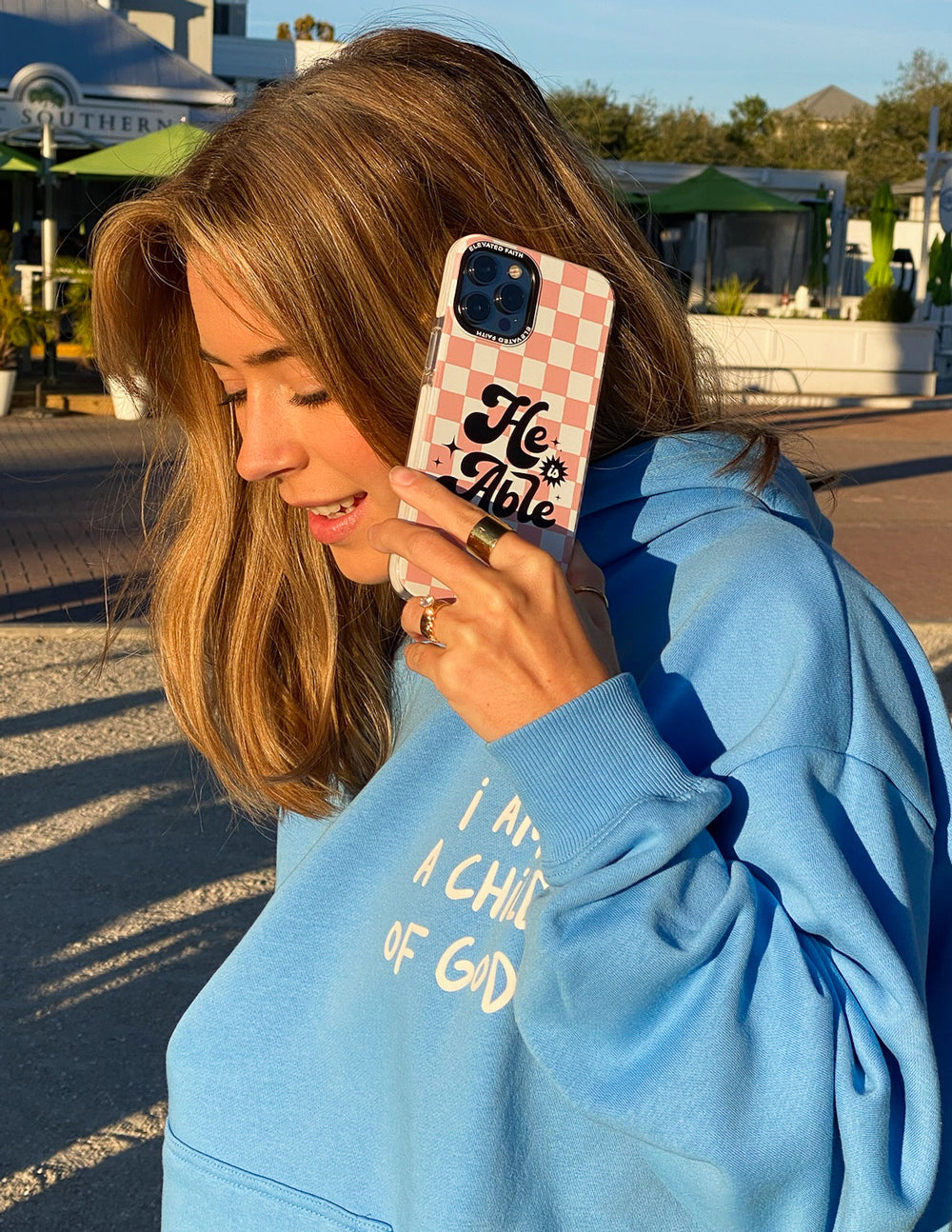 Pink checkered phone case with 'He is Able' on back, in use