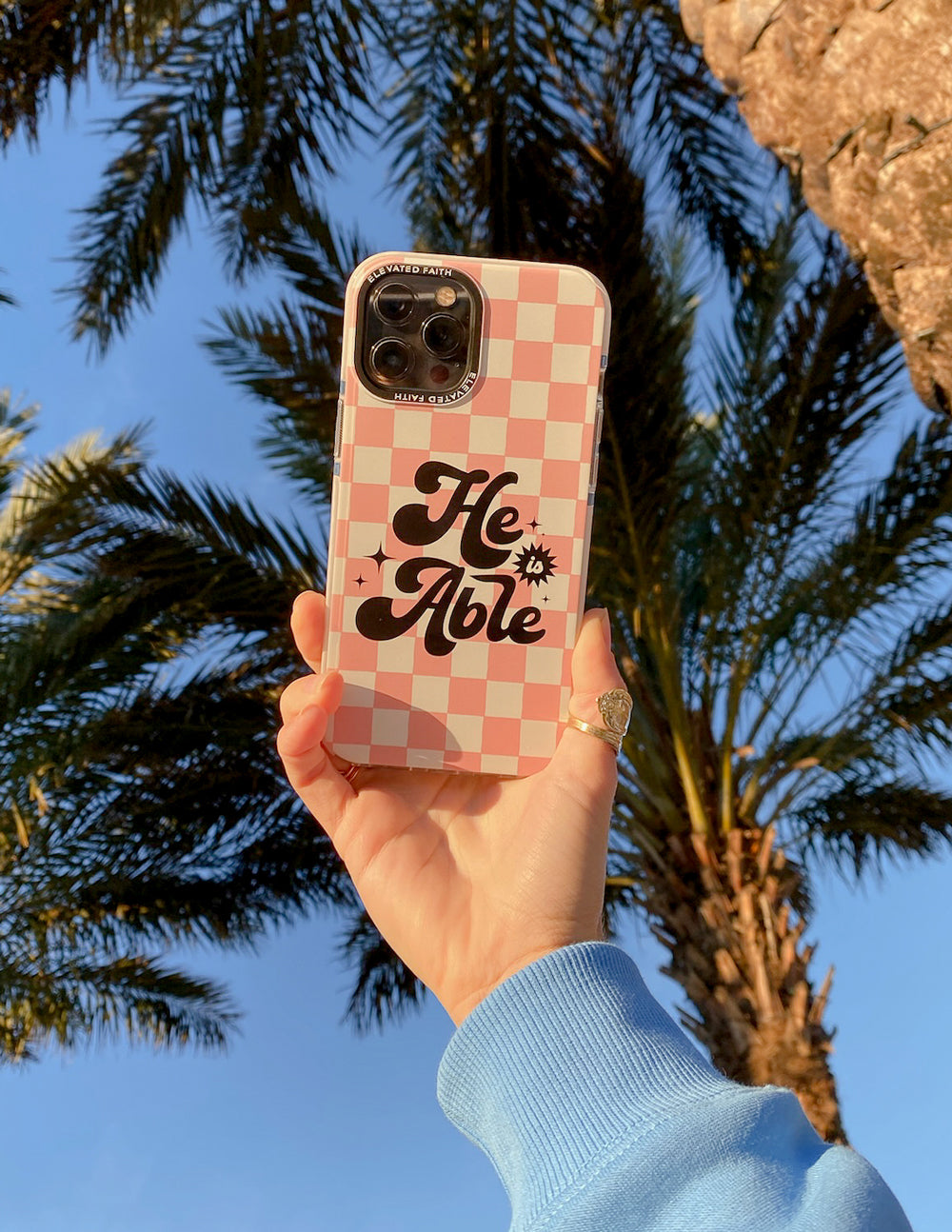 Pink checkered phone case with 'He is Able' logo, held upright