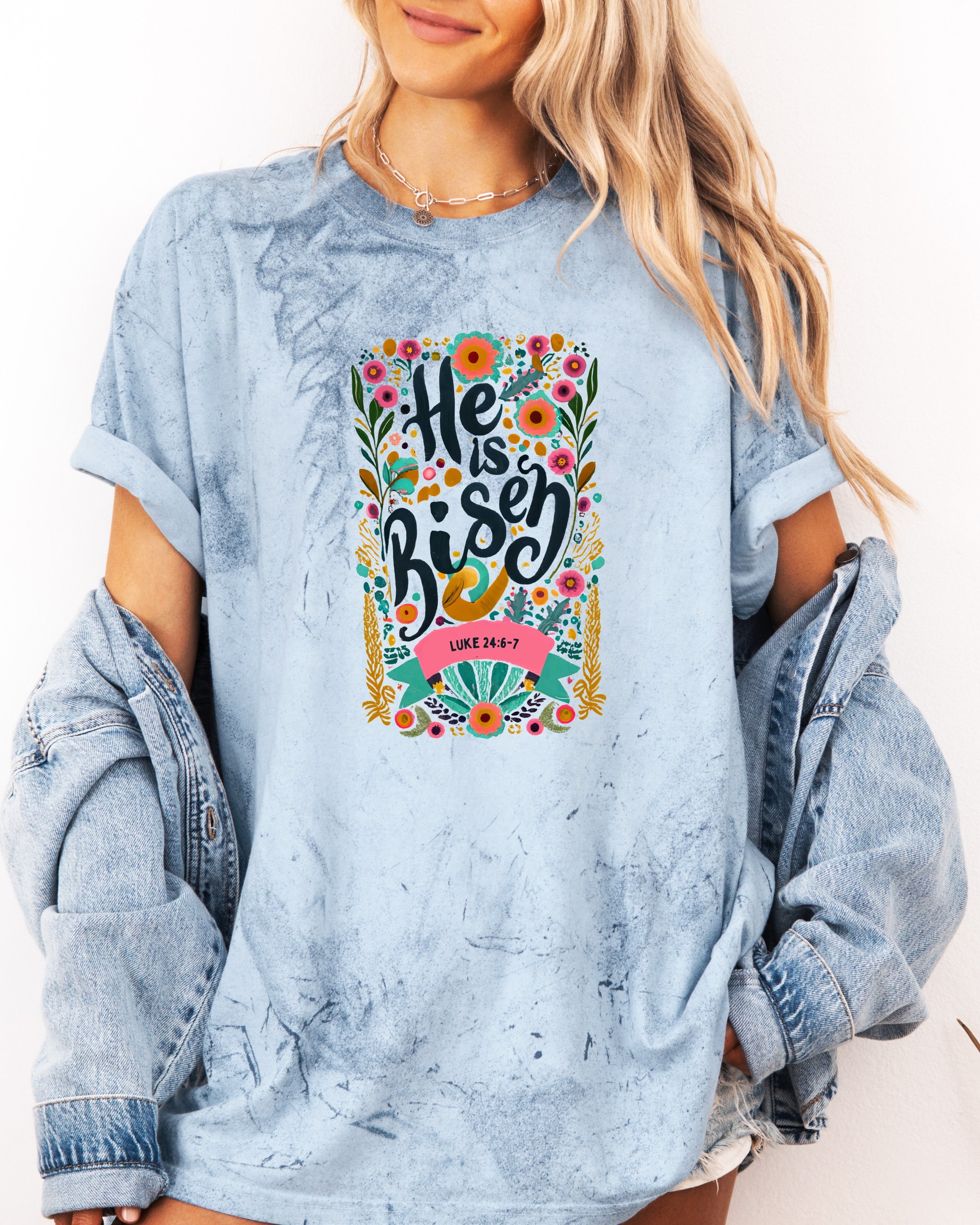 Garment-dyed light blue unisex T-shirt with colorful "He is Risen" front graphic