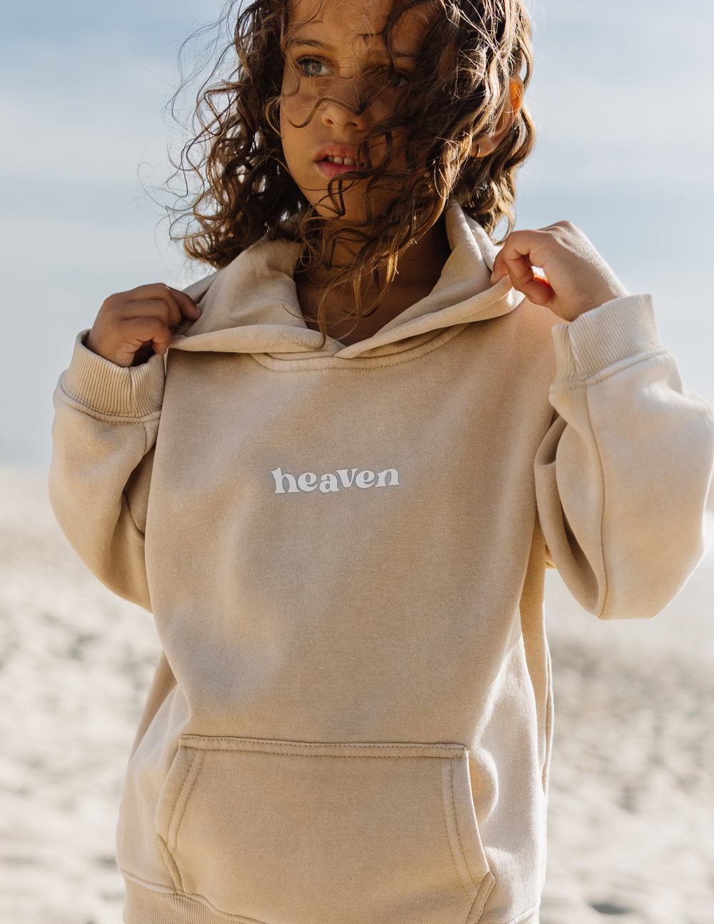 Kids hoodie in light tan with “heaven” logo on the chest and front pocket.