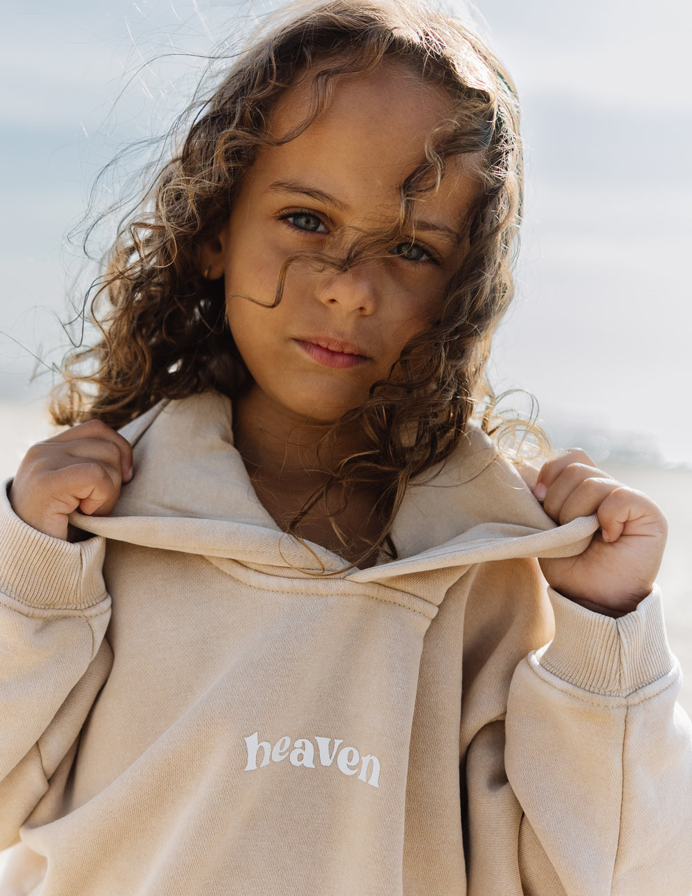 Kids hoodie in light tan with “heaven” logo on chest and drawstring hood.