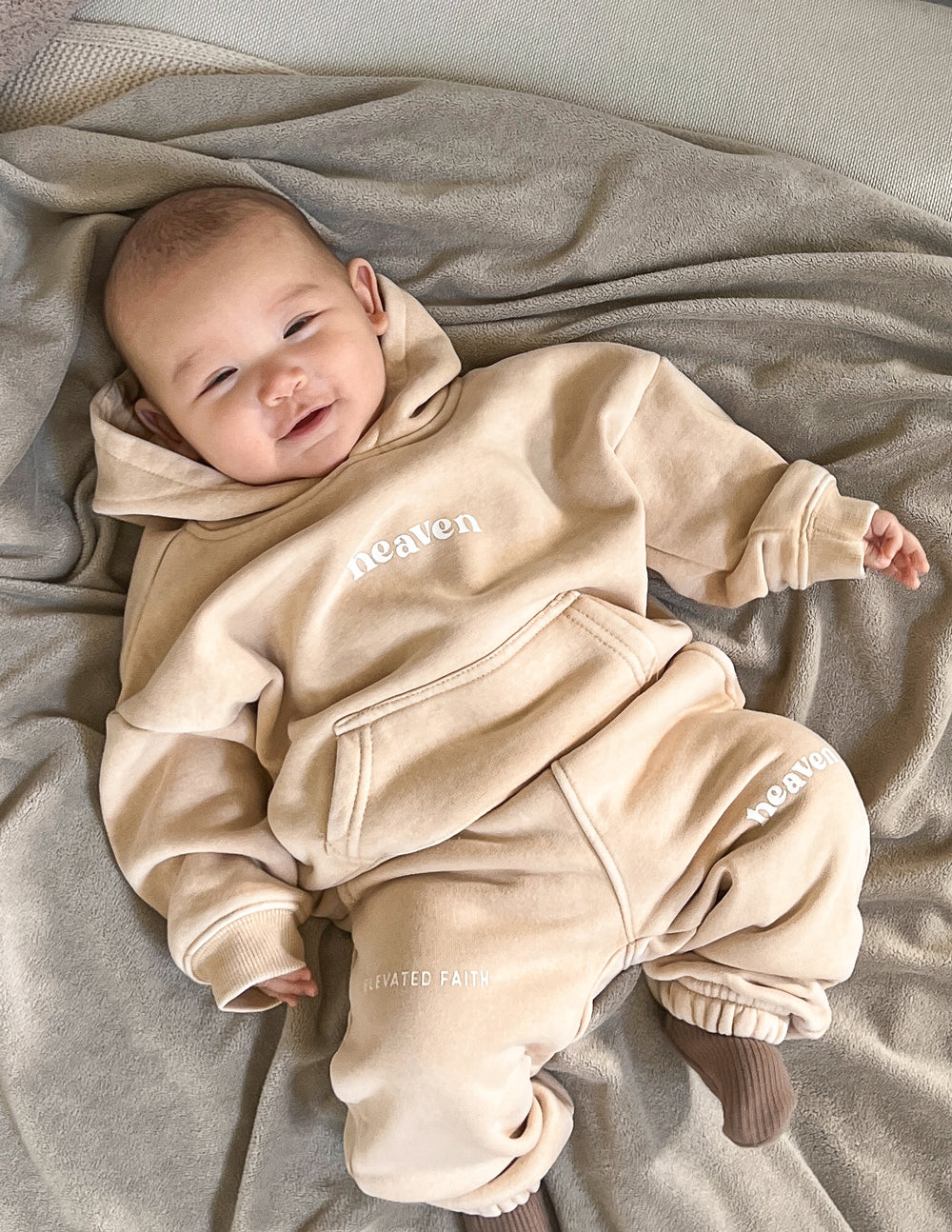 Baby hoodie in tan with “heaven” chest logo and front kangaroo pocket.