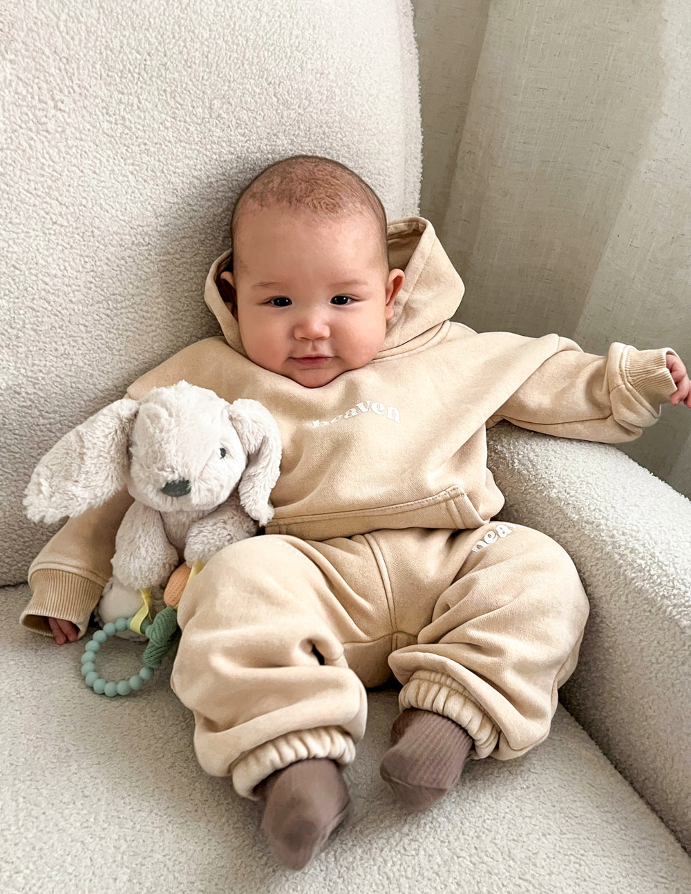Baby hoodie in tan with “heaven” logo, hood, and kangaroo pocket; long sleeves.