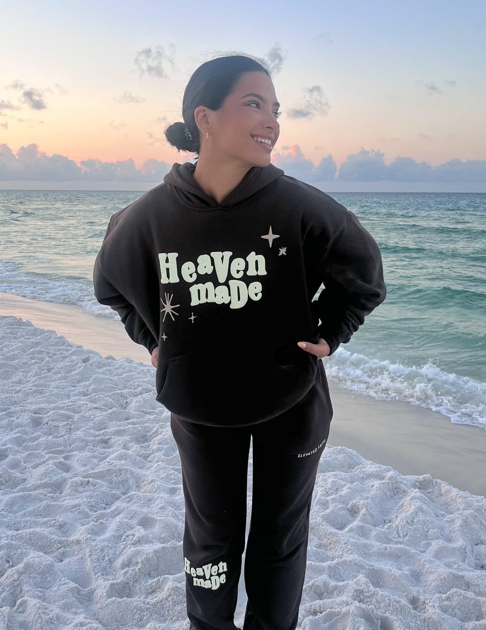 Dark brown unisex hoodie with front pocket and “Heaven Made” graphic; hands on hips.