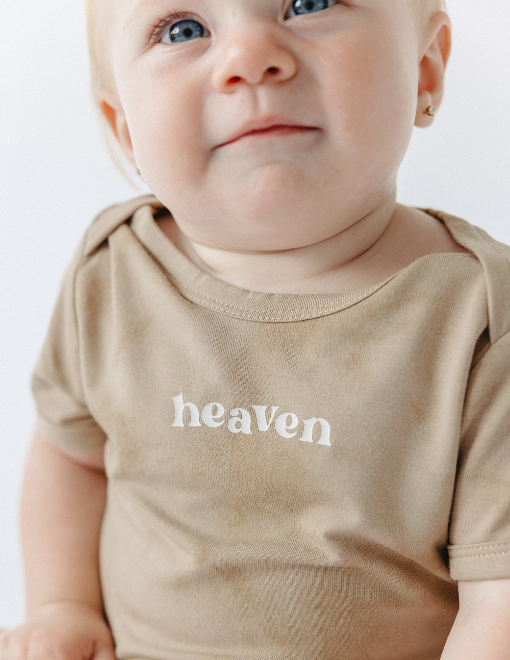 Tan baby onesie close-up with 'heaven' chest logo, short sleeves and crew neck
