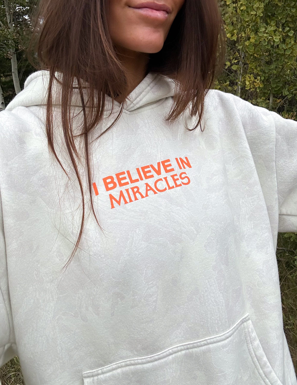 Close-up of light camo unisex hoodie with orange "I BELIEVE IN MIRACLES" chest print