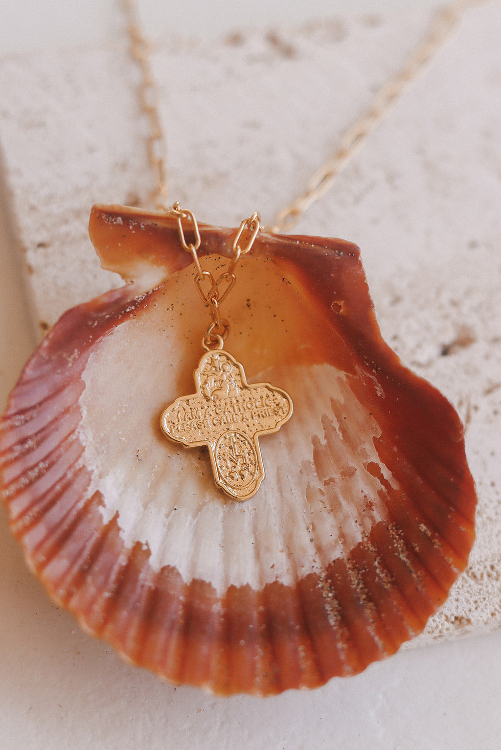 Gold paperclip chain necklace with cross pendant resting on seashell