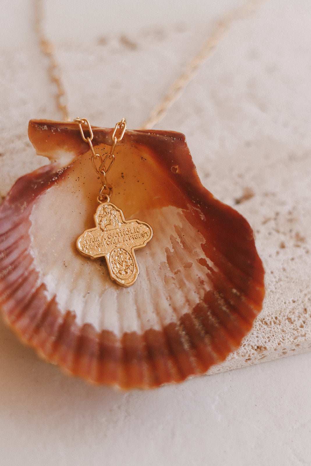 Gold cross pendant on paperclip chain necklace, resting on a shell
