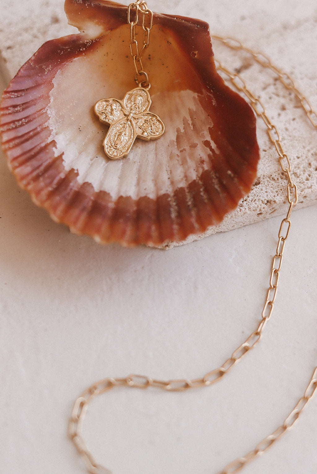 Gold cross pendant necklace with paperclip chain, shown on a shell
