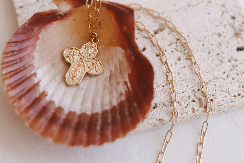 Gold cross pendant necklace with paperclip chain and clasp, shown on a shell