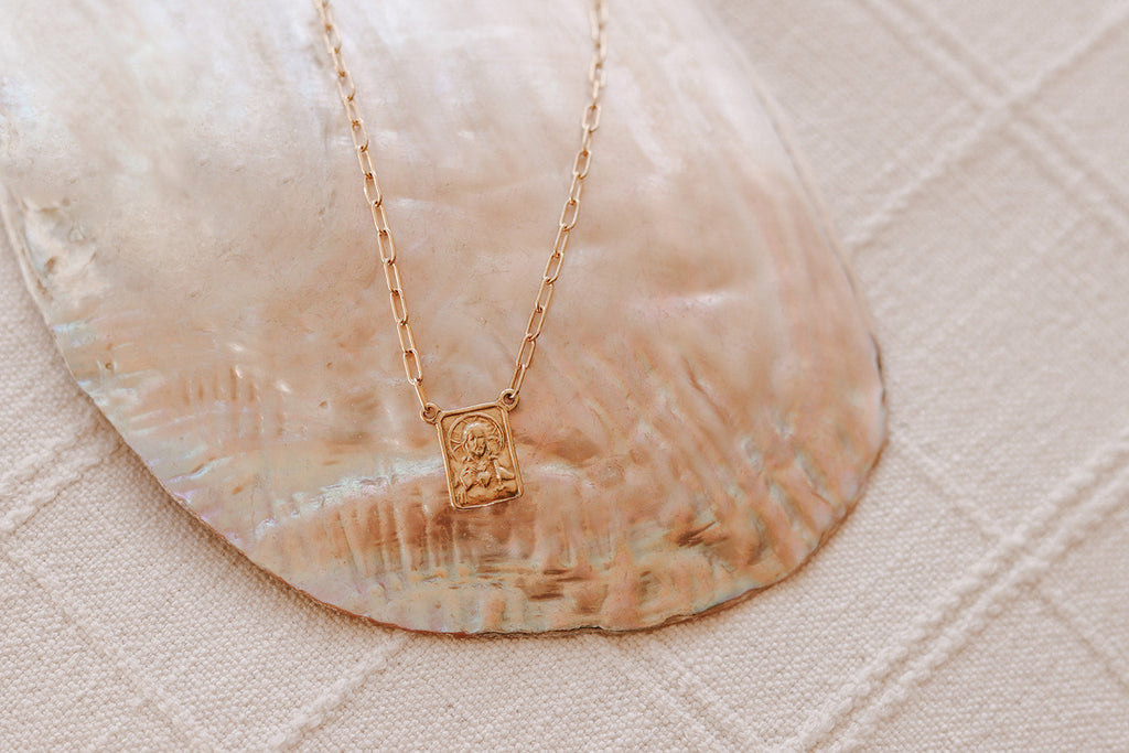 Scapular charm necklace with gold-tone rectangular pendant on light fabric